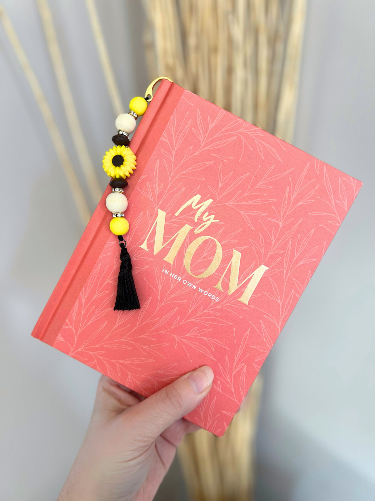 A handmade silicone beaded yellow sunflower daisy bookmark with rhinestones and wood accent beads finished with a chocolate brown faux silk fringe drop tassel laying over an inspirational journal book called "My Mom In Her Own Words"
