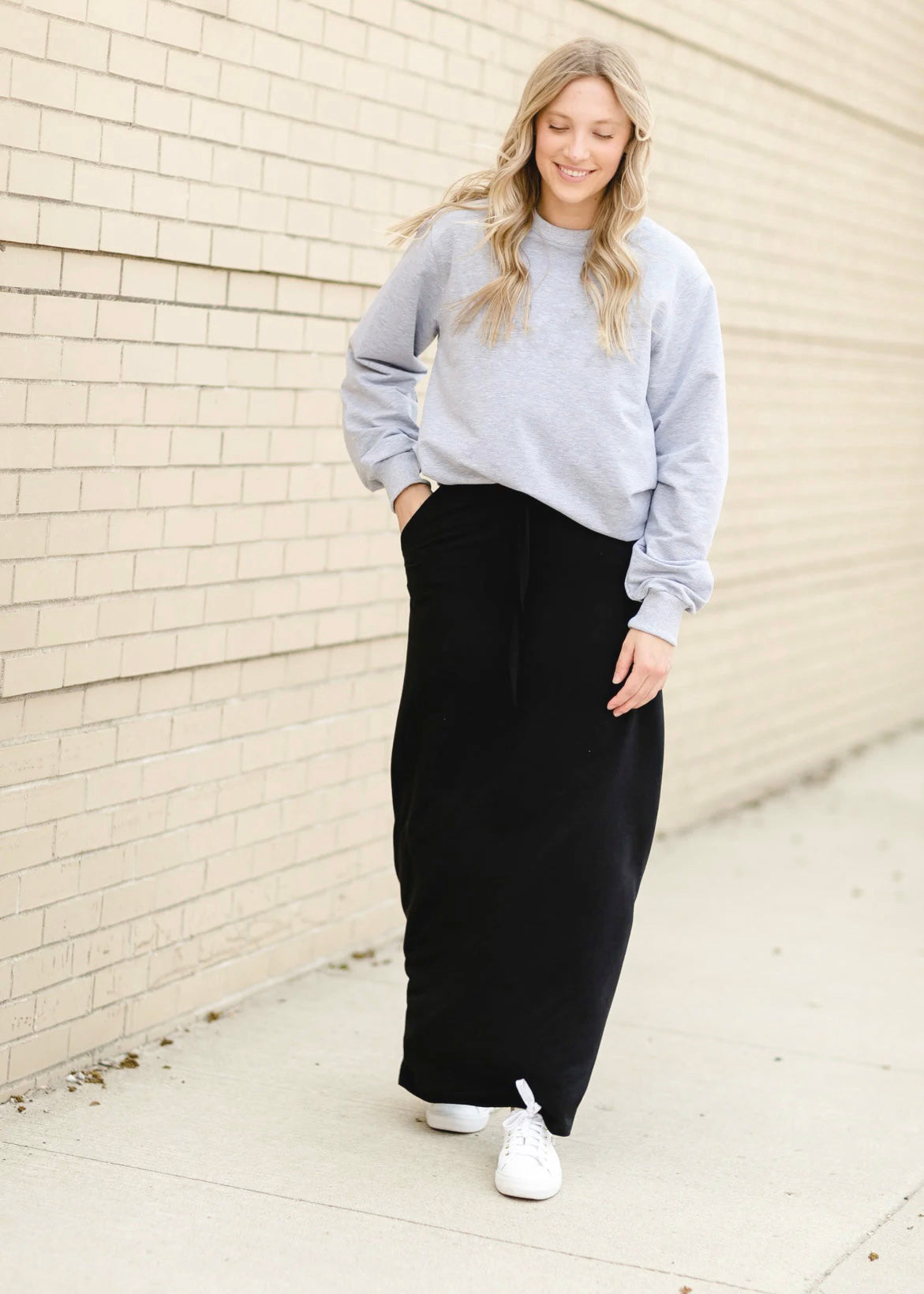 A young blonde woman with her hand in her pocket while smiling and wearing a sweatshirt and solid black Maxi Skirt with Pockets featuring a Soft & Stretchy Premium Fabric Blend, Adjustable Drawstring Tie Waist, and Pockets. Available in sizes S-3XL in both solid Black or Navy Blue. Discover our Best Selling All-year All-season Maxi Skirt for every body shape & size.