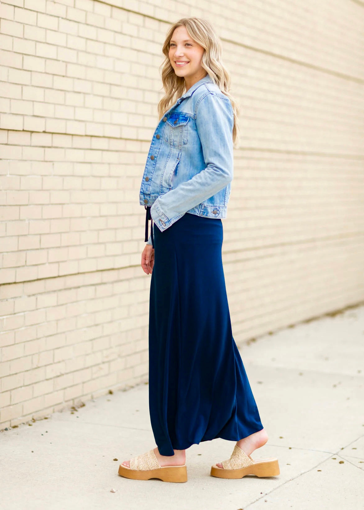 Side view of a young blonde woman wearing a denim jacket and navy blue Maxi Skirt with Pockets featuring a Soft & Stretchy Premium Fabric Blend, Adjustable Drawstring Tie Waist, and Pockets. Available in sizes S-3XL in both solid Black or Navy Blue. Discover our Best Selling All-year All-season Maxi Skirt for every body shape & size. 