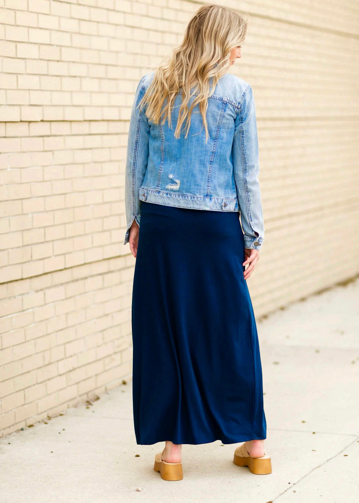 Rear view of a young blonde woman wearing a denim jacket and navy blue Maxi Skirt with Pockets featuring a Soft & Stretchy Premium Fabric Blend, Adjustable Drawstring Tie Waist, and Pockets. Available in sizes S-3XL in both solid Black or Navy Blue. Discover our Best Selling All-year All-season Maxi Skirt for every body shape & size. 