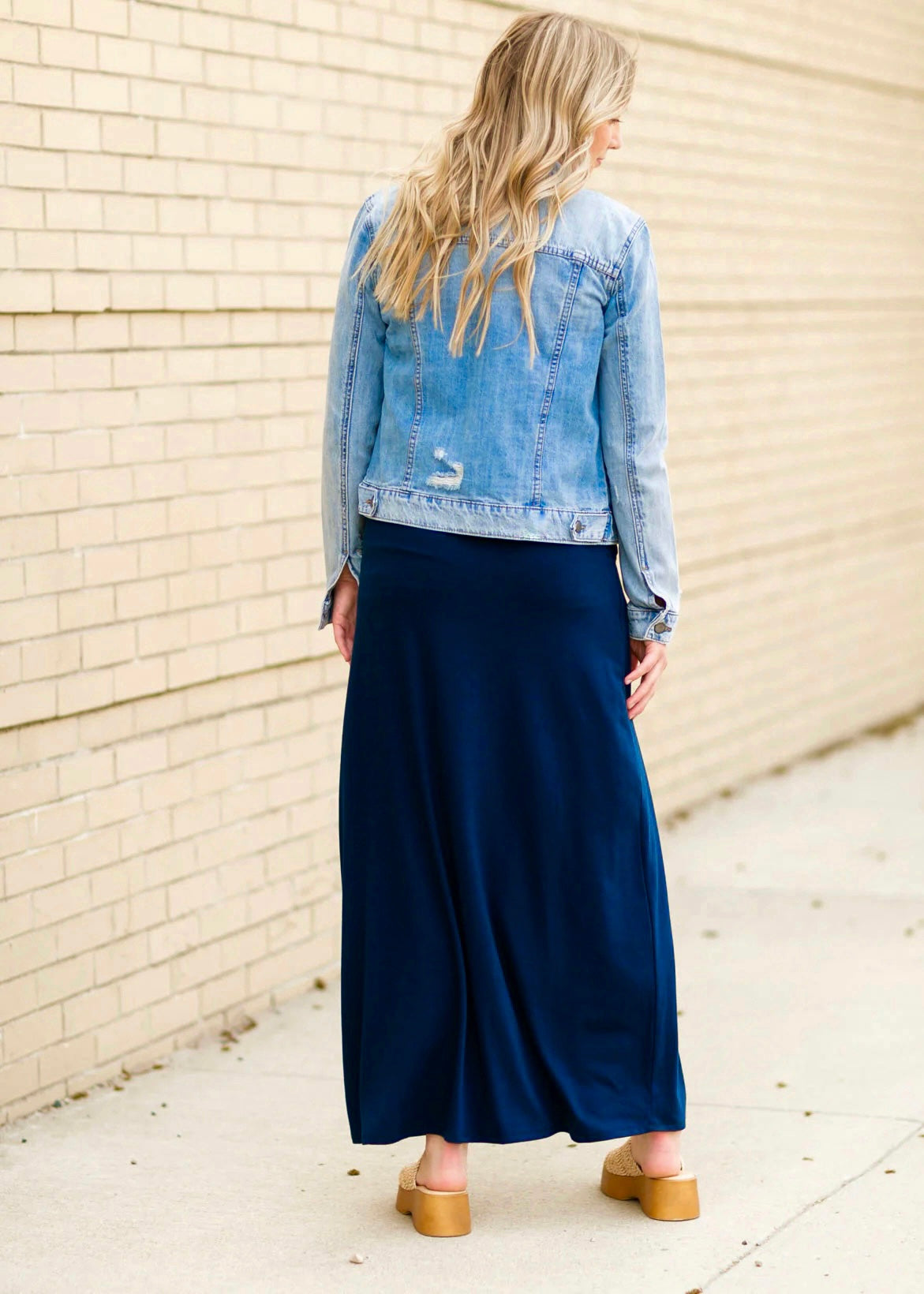 Rear view of a young blonde woman wearing a denim jacket and navy blue Maxi Skirt with Pockets featuring a Soft & Stretchy Premium Fabric Blend, Adjustable Drawstring Tie Waist, and Pockets. Available in sizes S-3XL in both solid Black or Navy Blue. Discover our Best Selling All-year All-season Maxi Skirt for every body shape & size.