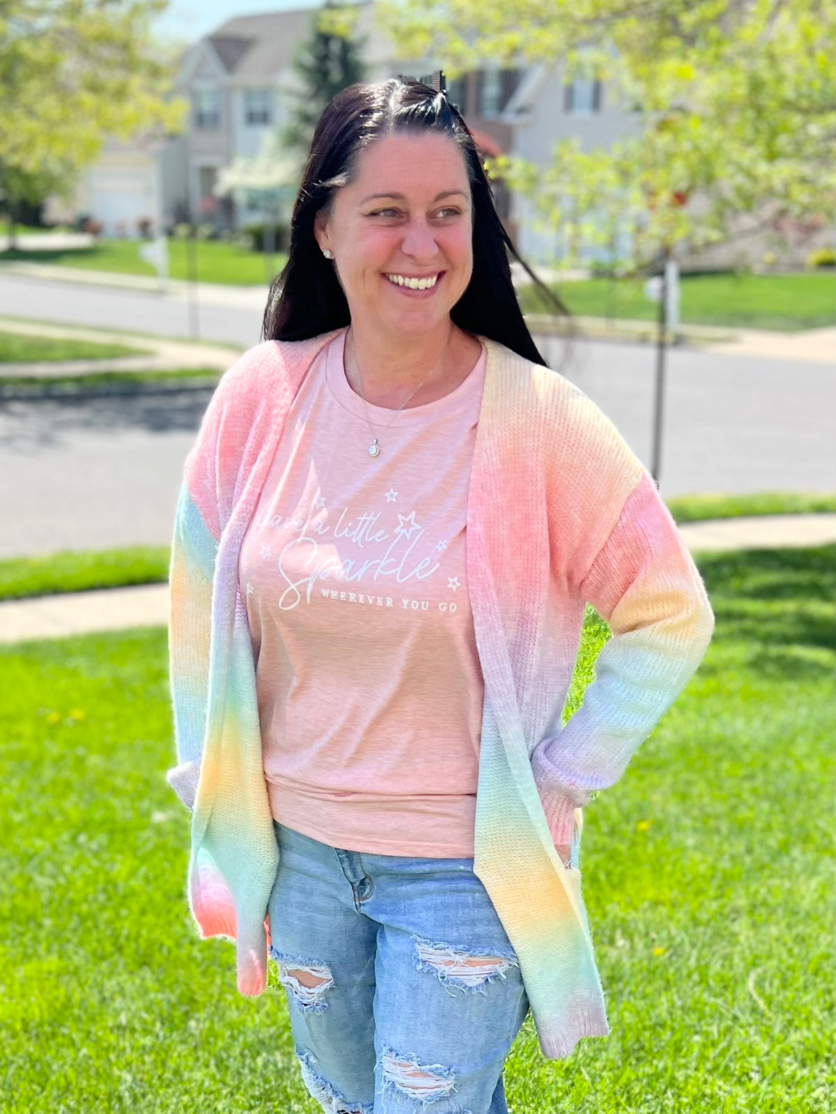 Woman outside wearing a rainbow ombre cardigan, leave a little sparkle graphic t-shirt, judy blue jeans, and moissanite solitaire earrings and necklace