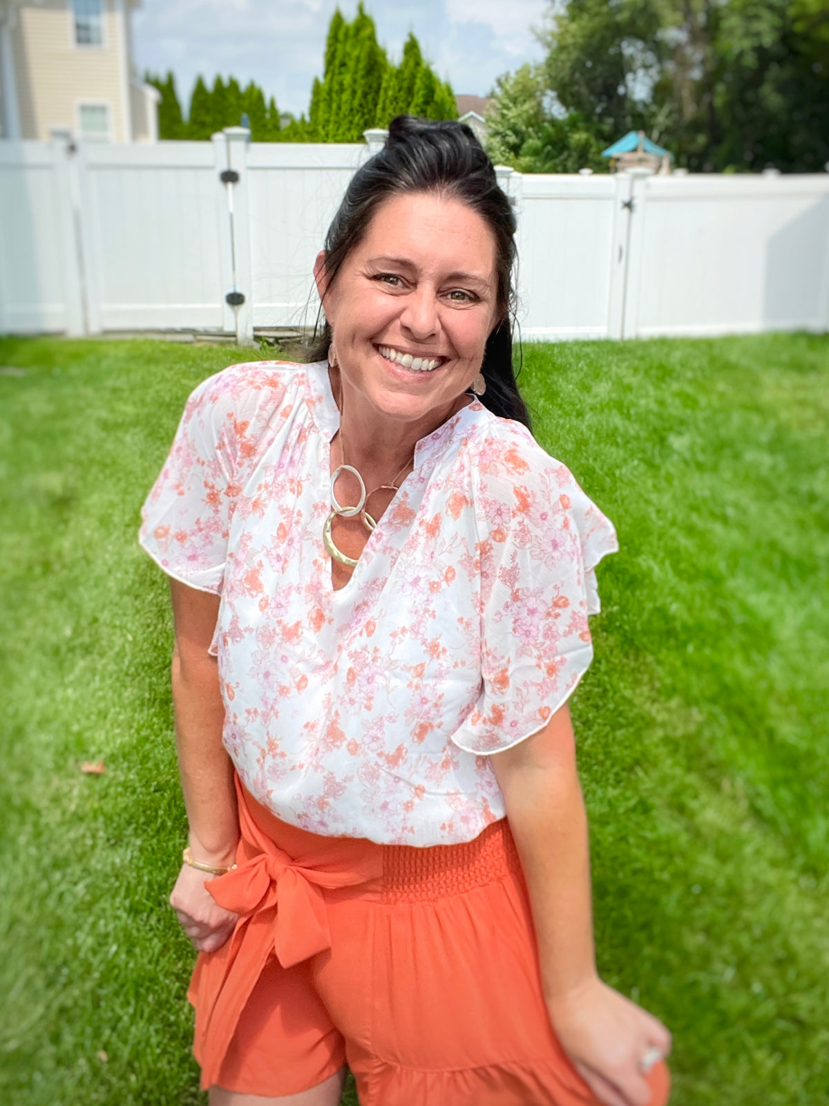 Woman smiling standing outside on a sunny day wearing a floral flutter sleeve notched neck blouse with smocked tie waist terracotta orange swing shorts and a hammered oval necklace with druzy open stone earrings