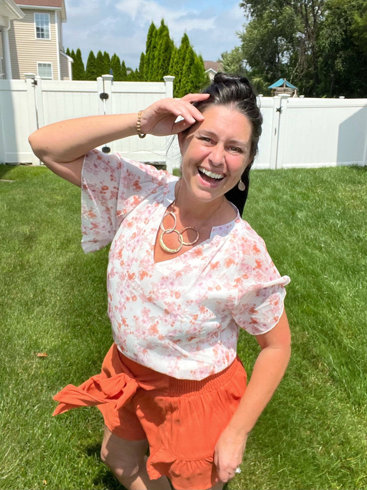 Brunette Woman laughing with one hand on her forehead holding back her hair while the wind in blowing. She is wearing a floral flutter sleeve notched neck blouse with smocked tie waist terracotta orange swing shorts and a hammered oval necklace with druzy open stone earrings