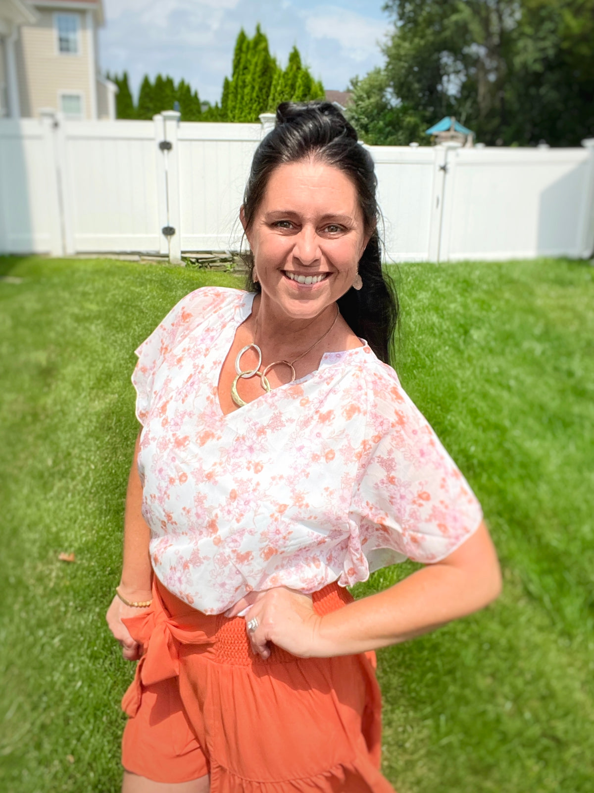 Woman smiling posing for the camera wearing a floral flutter sleeve notched neck blouse with smocked tie waist terracotta orange swing shorts and a hammered oval necklace with druzy open stone earrings