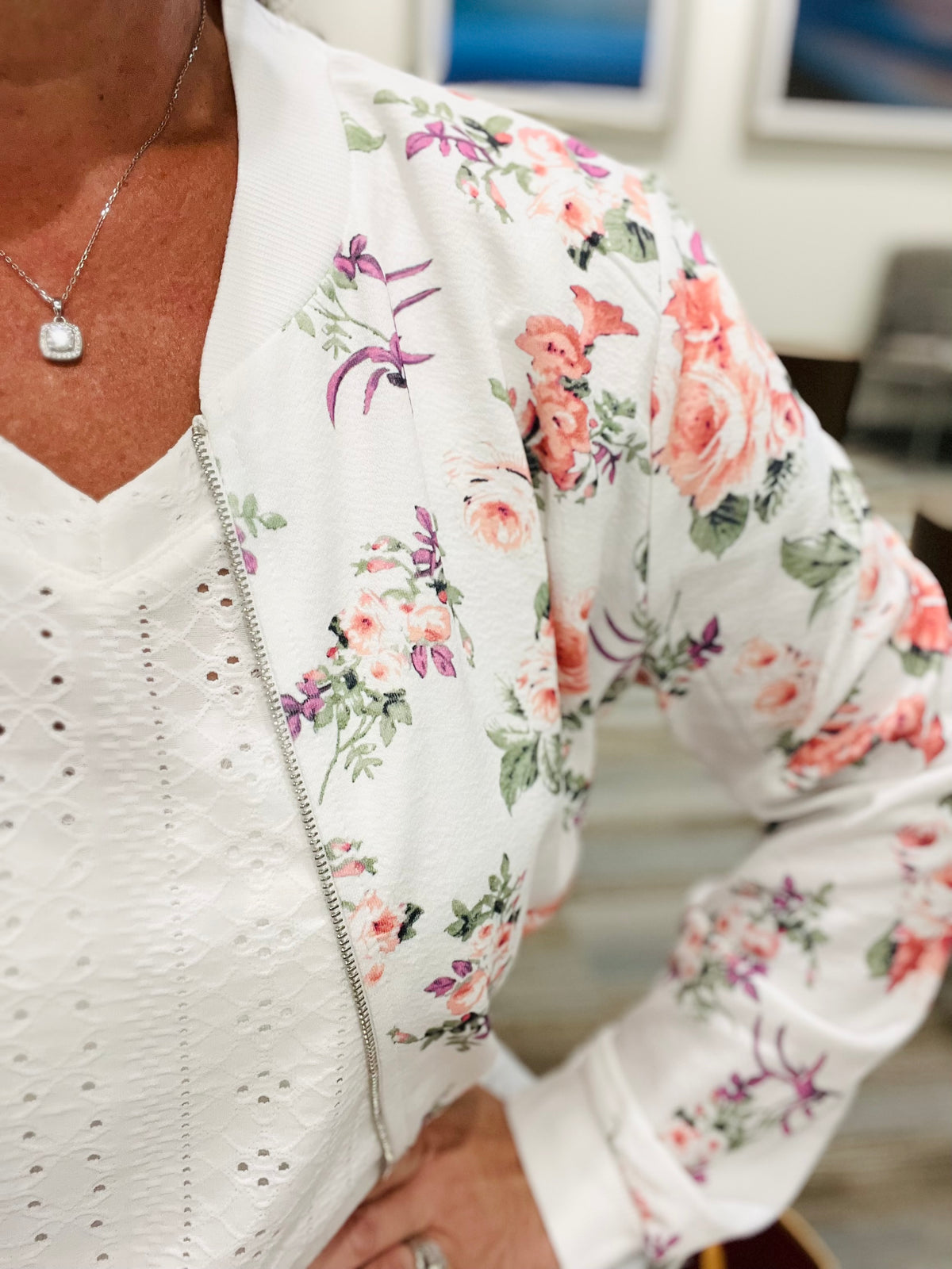 A close up view of a woman's arm wearing a white floral zip up lightweight floral bomber jacket unzipped over a white eyelet v-neck top paired with a solitaire moissanite necklace