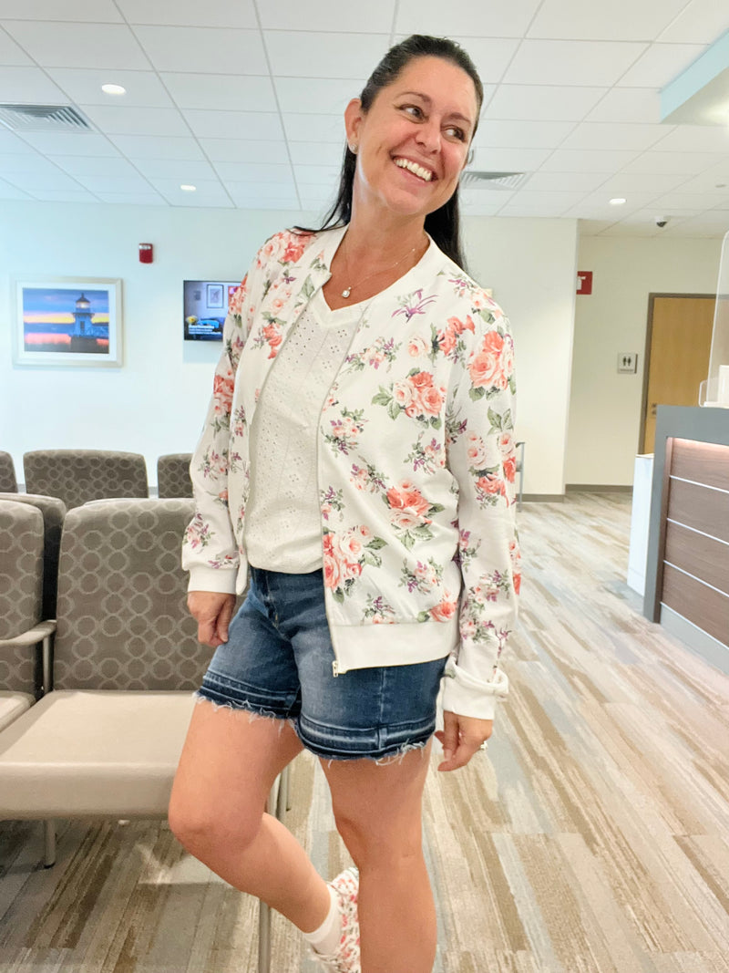 Brunette Woman standing & smiling while looking over her shoulder wearing a white floral zip up lightweight floral bomber jacket unzipped over a white eyelet v-neck top paired with denim shorts and floral sneakers