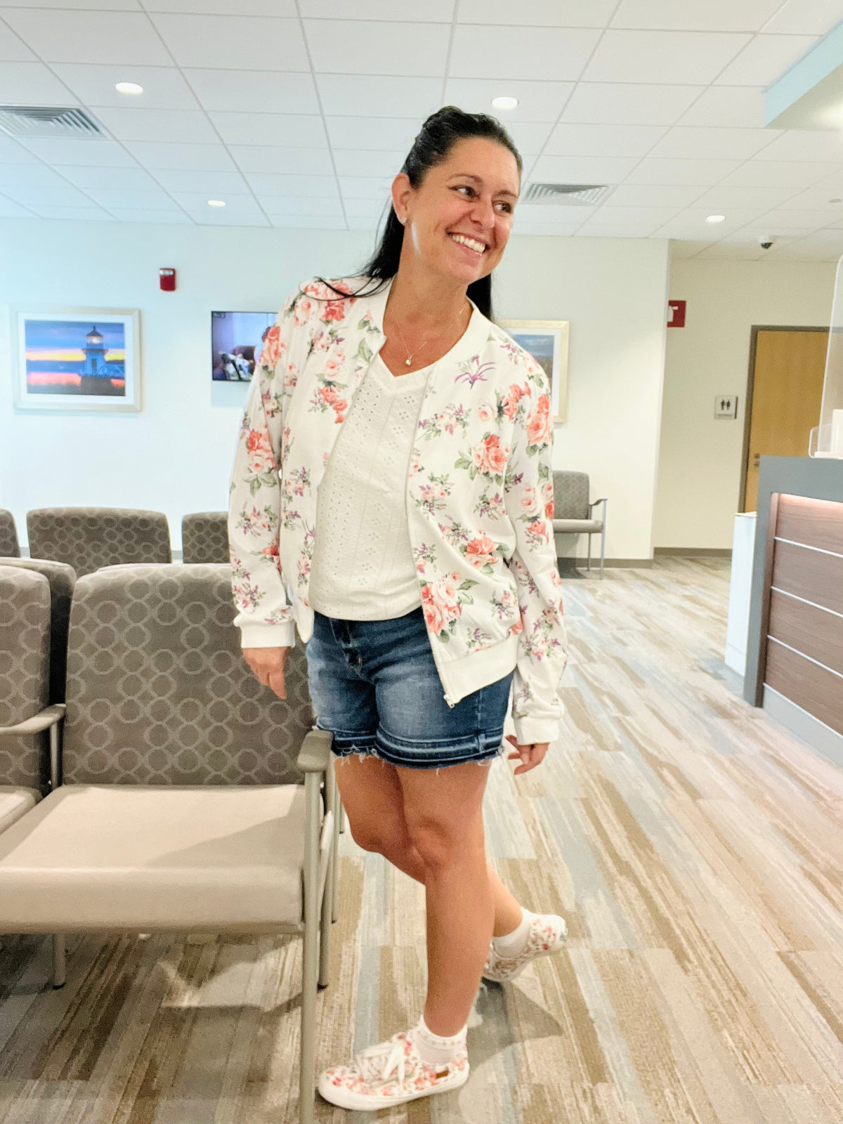 Brunette Woman standing & smiling while looking over her shoulder wearing a white floral zip up lightweight floral bomber jacket unzipped over a white eyelet v-neck top paired with denim shorts and floral sneakers