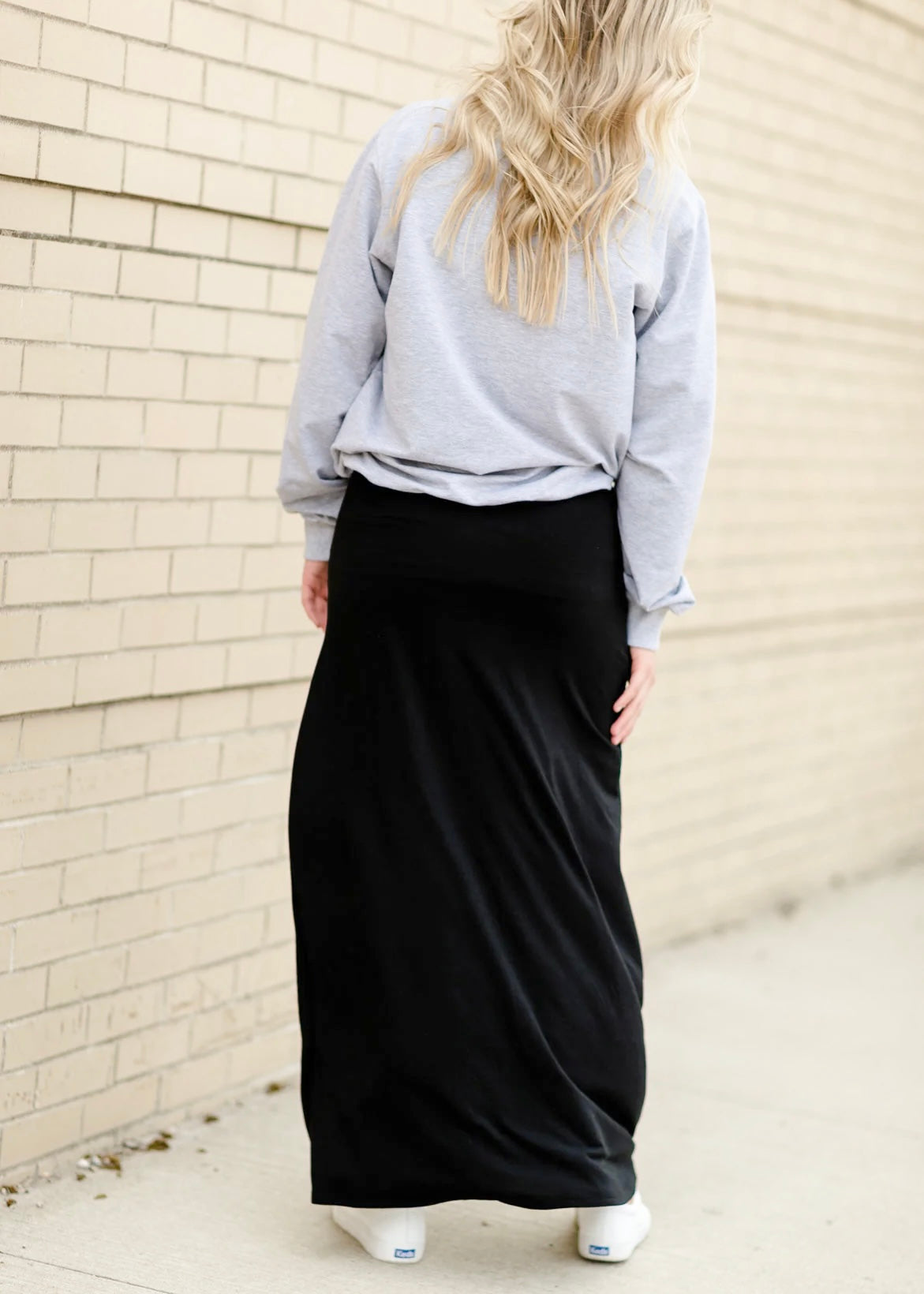 Rear view of a young woman wearing a solid black Maxi Skirt with Pockets featuring a Soft & Stretchy Premium Fabric Blend, Adjustable Drawstring Tie Waist, and Pockets. Available in sizes S-3XL in both solid Black or Navy Blue. Discover our Best Selling All-year All-season Maxi Skirt for every body shape & size.