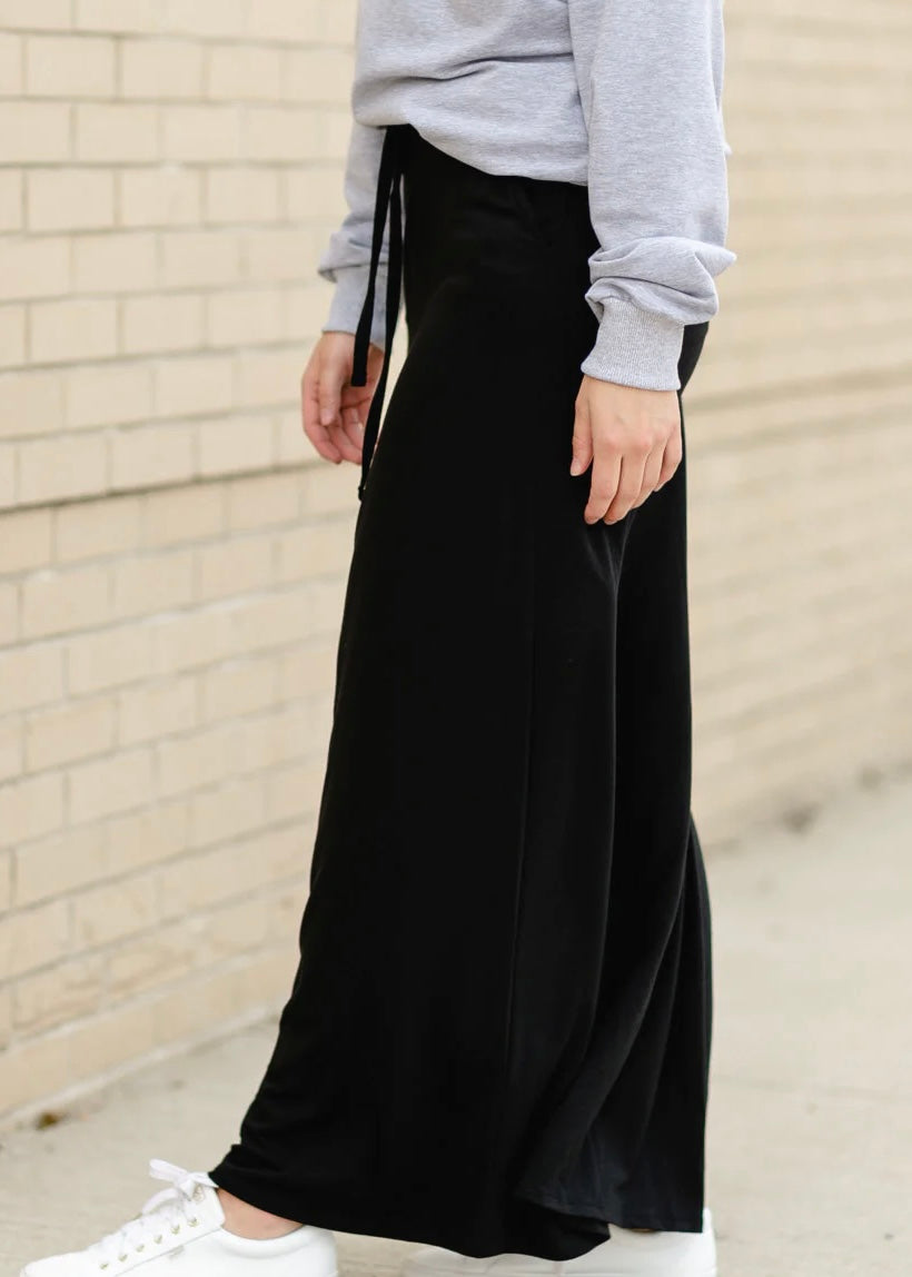 Bottom half side view of a young woman wearing a solid black Maxi Skirt with Pockets featuring a Soft & Stretchy Premium Fabric Blend, Adjustable Drawstring Tie Waist, and Pockets. Available in sizes S-3XL in both solid Black or Navy Blue. Discover our Best Selling All-year All-season Maxi Skirt for every body shape & size.