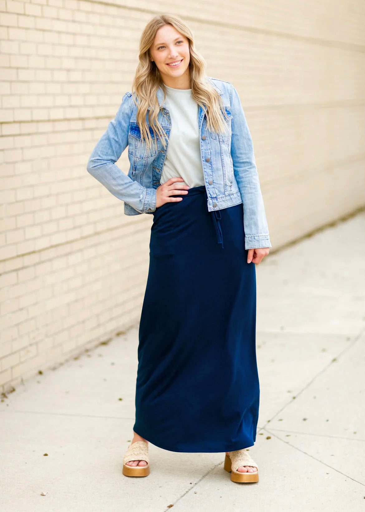 A young blonde woman with her hand on her hip smiling and wearing a denim jacket and navy blue Maxi Skirt with Pockets featuring a Soft & Stretchy Premium Fabric Blend, Adjustable Drawstring Tie Waist, and Pockets. Available in sizes S-3XL in both solid Black or Navy Blue. Discover our Best Selling All-year All-season Maxi Skirt for every body shape & size. 