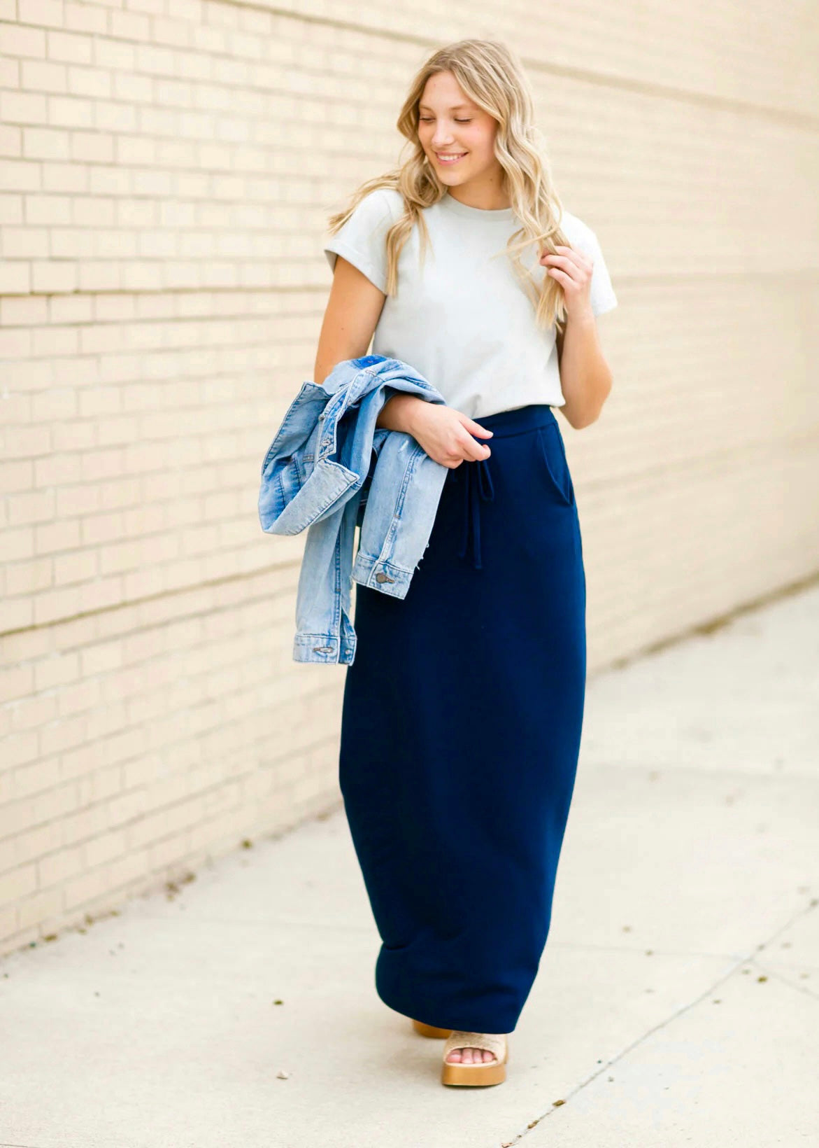 A young blonde woman holding a denim jacket in her arm while smiling and wearing a navy blue Maxi Skirt with Pockets featuring a Soft & Stretchy Premium Fabric Blend, Adjustable Drawstring Tie Waist, and Pockets. Available in sizes S-3XL in both solid Black or Navy Blue. Discover our Best Selling All-year All-season Maxi Skirt for every body shape & size. 