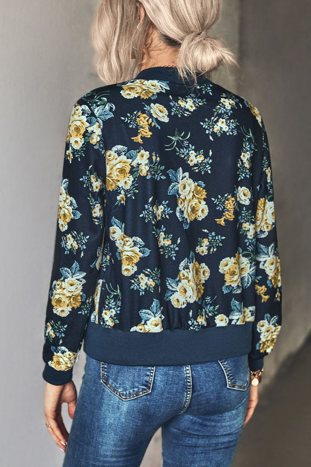 Back view of a Blonde Woman wearing a navy blue floral zip up lightweight floral bomber jacket unzipped over a white tube top paired with belted denim blue jeans