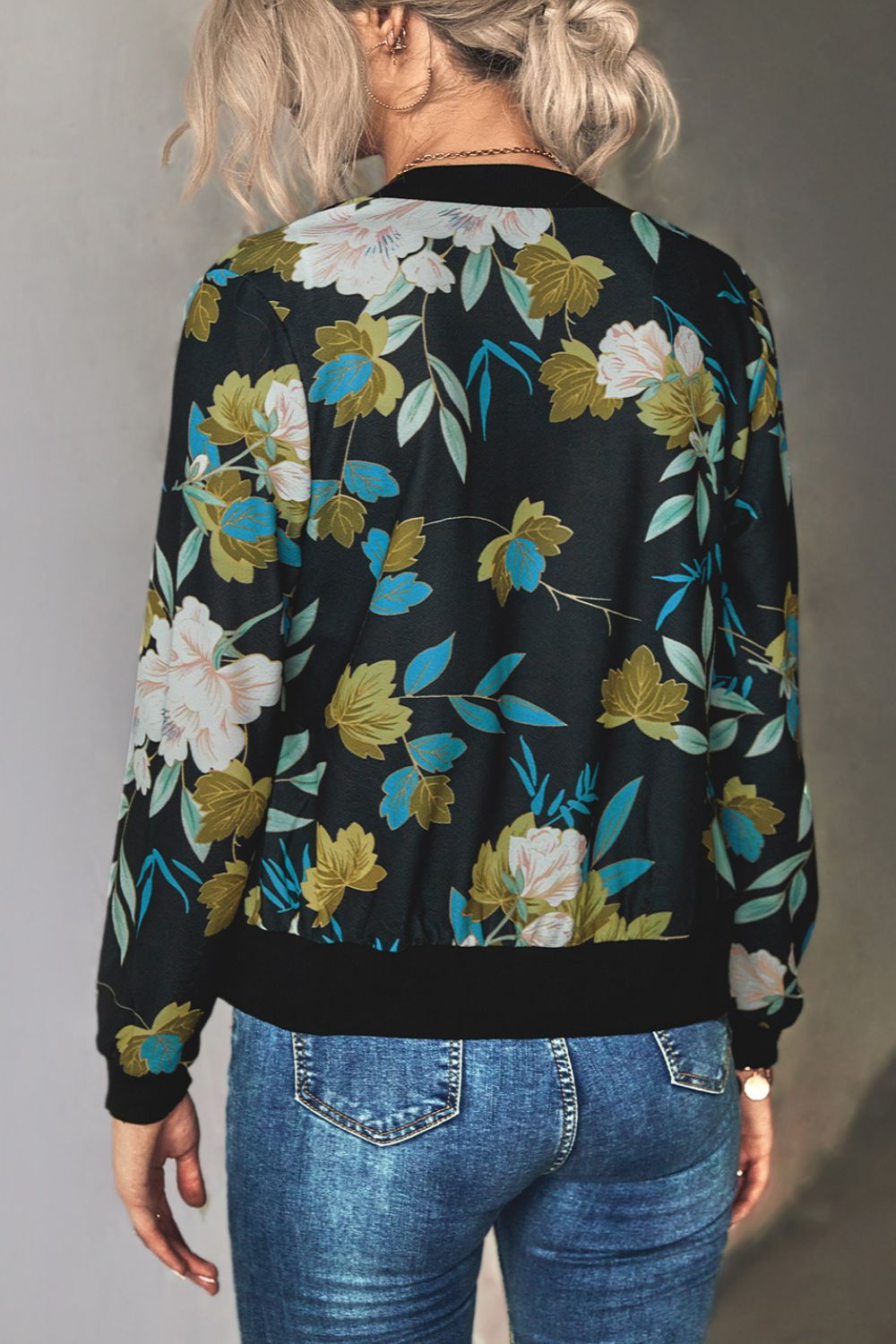 The back view of a Woman wearing a black floral zip up lightweight floral bomber jacket with jeans