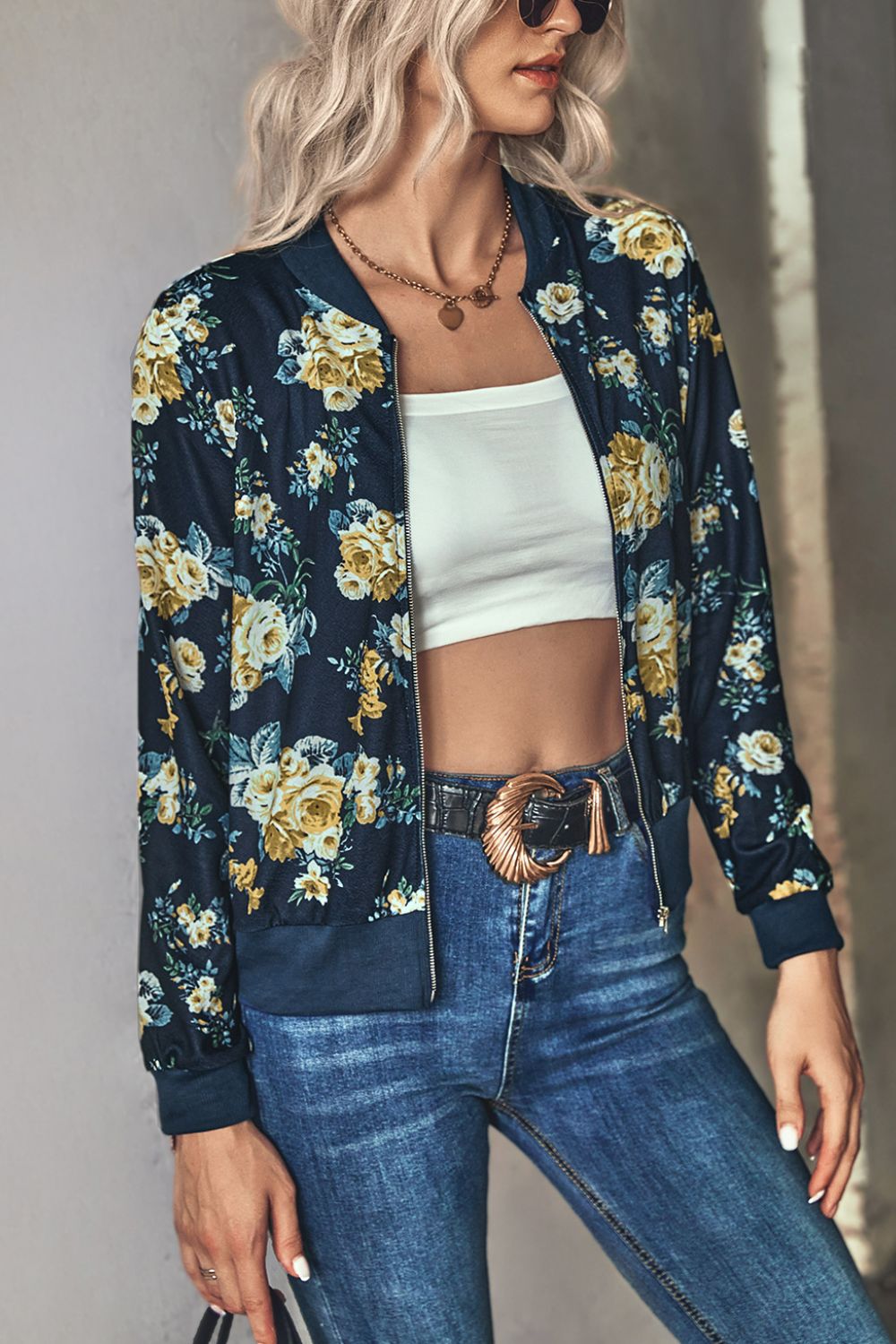 Blonde Woman wearing a navy blue floral zip up lightweight floral bomber jacket unzipped over a white tube top paired with belted denim blue jeans