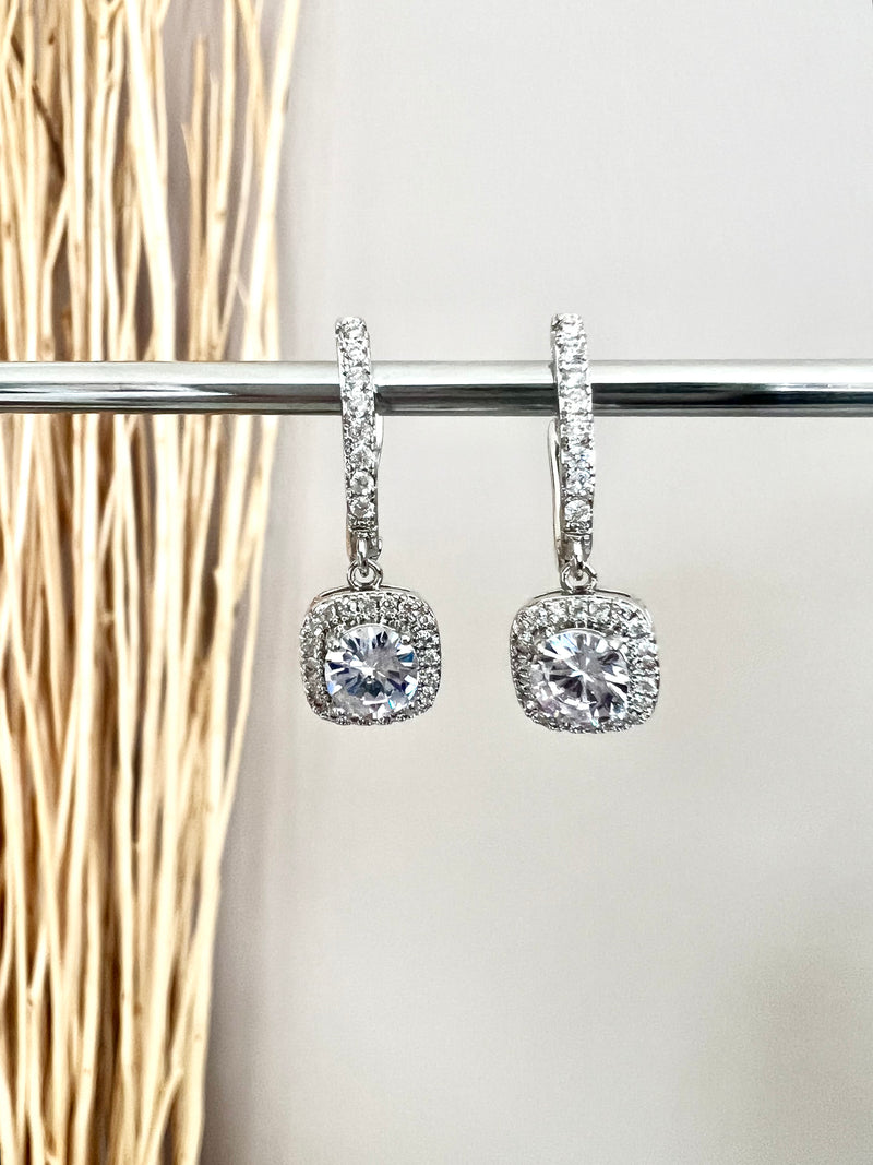 The Eleanor English Lock Embraced Solitaire Drop Earrings are crafted with a round solitaire in a square setting, secured by an English Lock Clasp for a secure fit. These earrings provide a timeless look with plenty of sparkle.   Material: 925 sterling silver rhodium-plated  Solitaire Cut: Brilliant Round  Setting: Halo Square Cushion  Length: 1"  Each earring: 1 carat  Total: 2 carat  Weight: 0.2 oz (6 g) for set