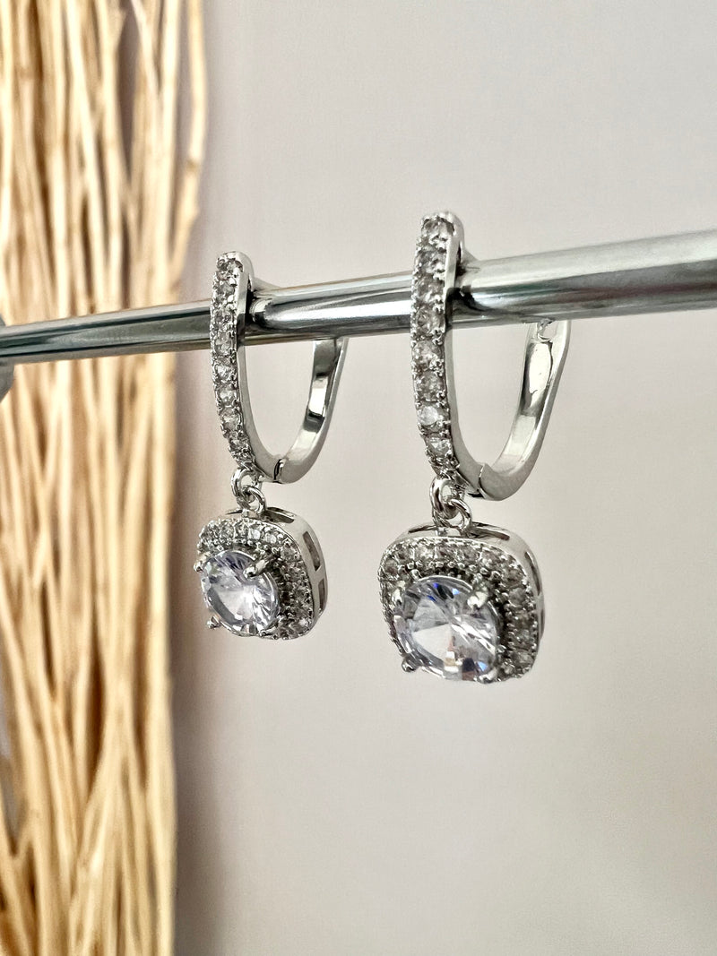 The Eleanor English Lock Embraced Solitaire Drop Earrings are crafted with a round solitaire in a square setting, secured by an English Lock Clasp for a secure fit. These earrings provide a timeless look with plenty of sparkle.   Material: 925 sterling silver rhodium-plated  Solitaire Cut: Brilliant Round  Setting: Halo Square Cushion  Length: 1"  Each earring: 1 carat  Total: 2 carat  Weight: 0.2 oz (6 g) for set